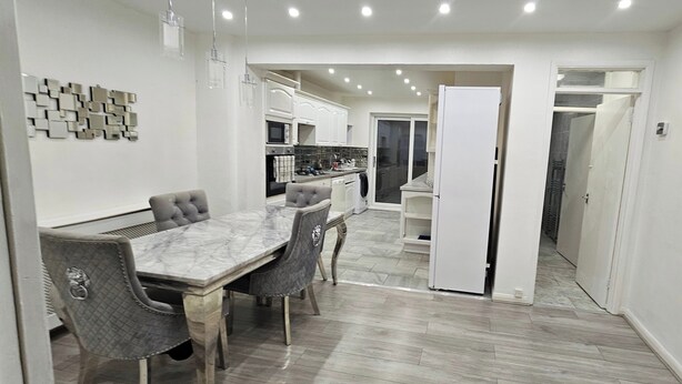 A modern kitchen with white cabinets, a marble countertop, and a dining area with a marble table and upholstered chairs. - Spacious & Modern 2-bed House in Dagenham (Dagenham)
