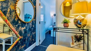 Een gang met een blauw-gouden spiegel, een trap, een bed en een tafel met een lamp en planten.