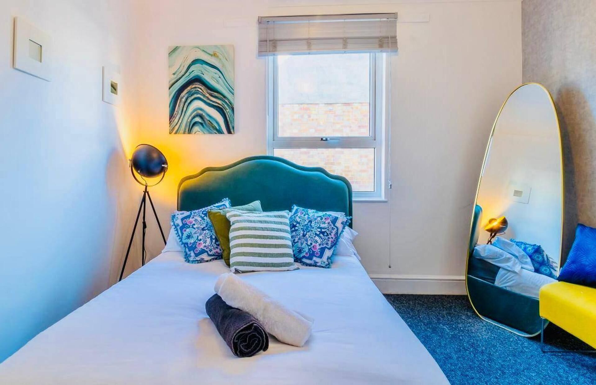A bedroom with a bed, a bedside lamp, a mirror, and a window.