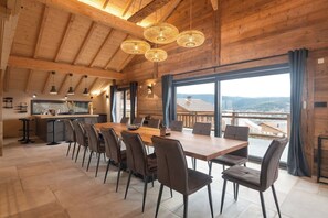 A spacious dining area with a long wooden table and modern chairs, a kitchen with a bar area, and large windows offering a view of the landscape.