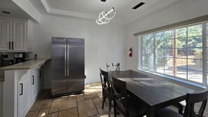 A modern kitchen with white cabinets, a stainless steel refrigerator, a dining table with chairs, and a large window providing a view of the outdoors. - Munguia Summit Camp|Modern 2BR·Near to Snow Summit (Big Bear Lake)