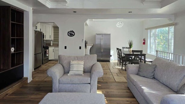 A spacious living room with a grey sofa, a coffee table, and a dining area with a table and chairs. A ceiling fan is visible, and there is a kitchen area with cabinets and appliances in the background. - Munguia Summit Camp|Modern 2BR·Near to Snow Summit (Big Bear Lake)