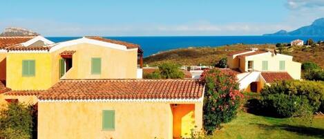 A coastal village with terracotta-roofed houses, a grassy area, and a view of the sea and mountains.