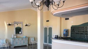 A hotel lobby with a reception desk, a mirror with a decorative frame, a chandelier, and a seating area with chairs. - Hotiday Golfo Aranci (Golfo Aranci)