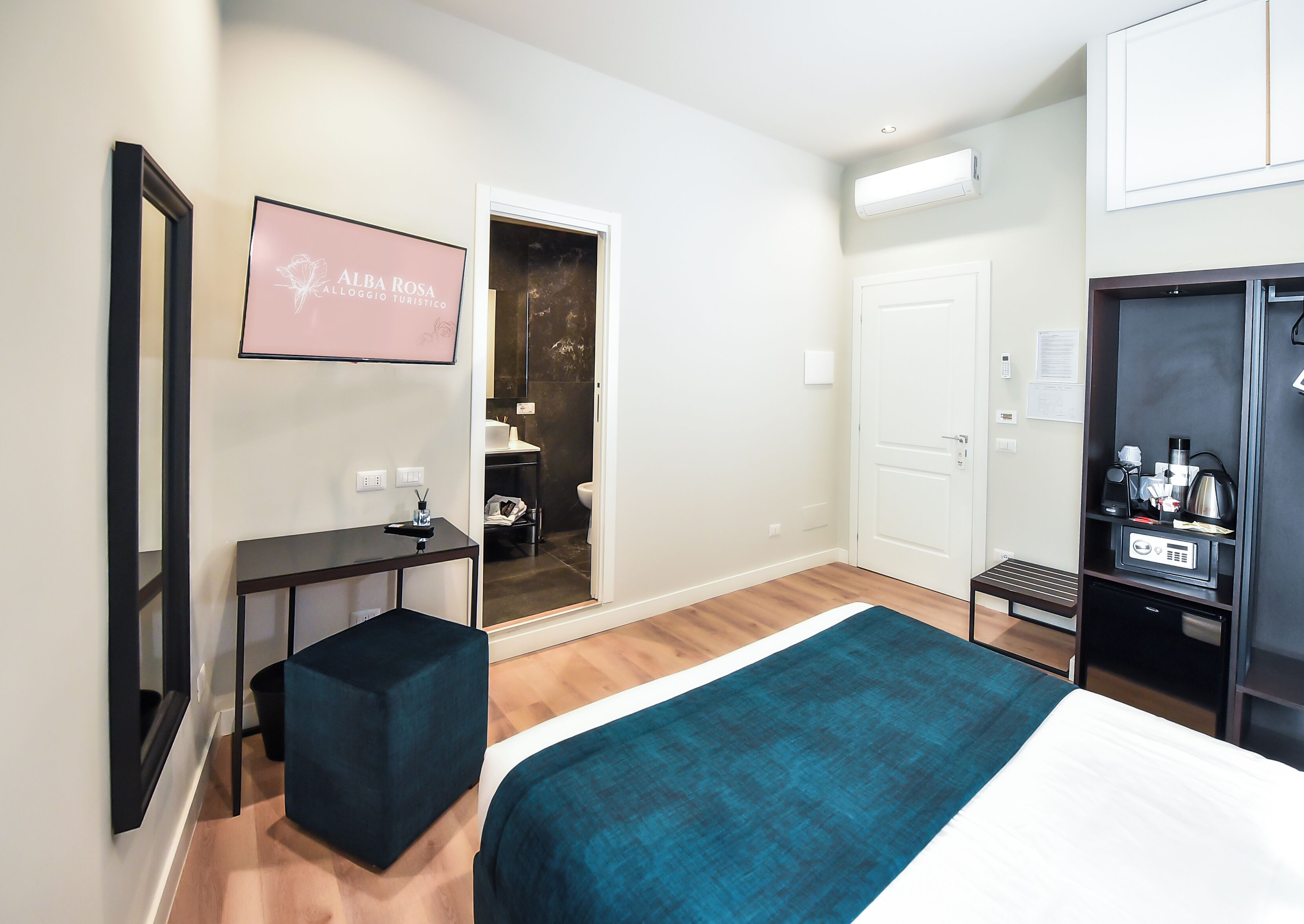 A hotel room with a bed, a desk, a mirror, a TV, a wardrobe, and a bathroom visible through an open door.