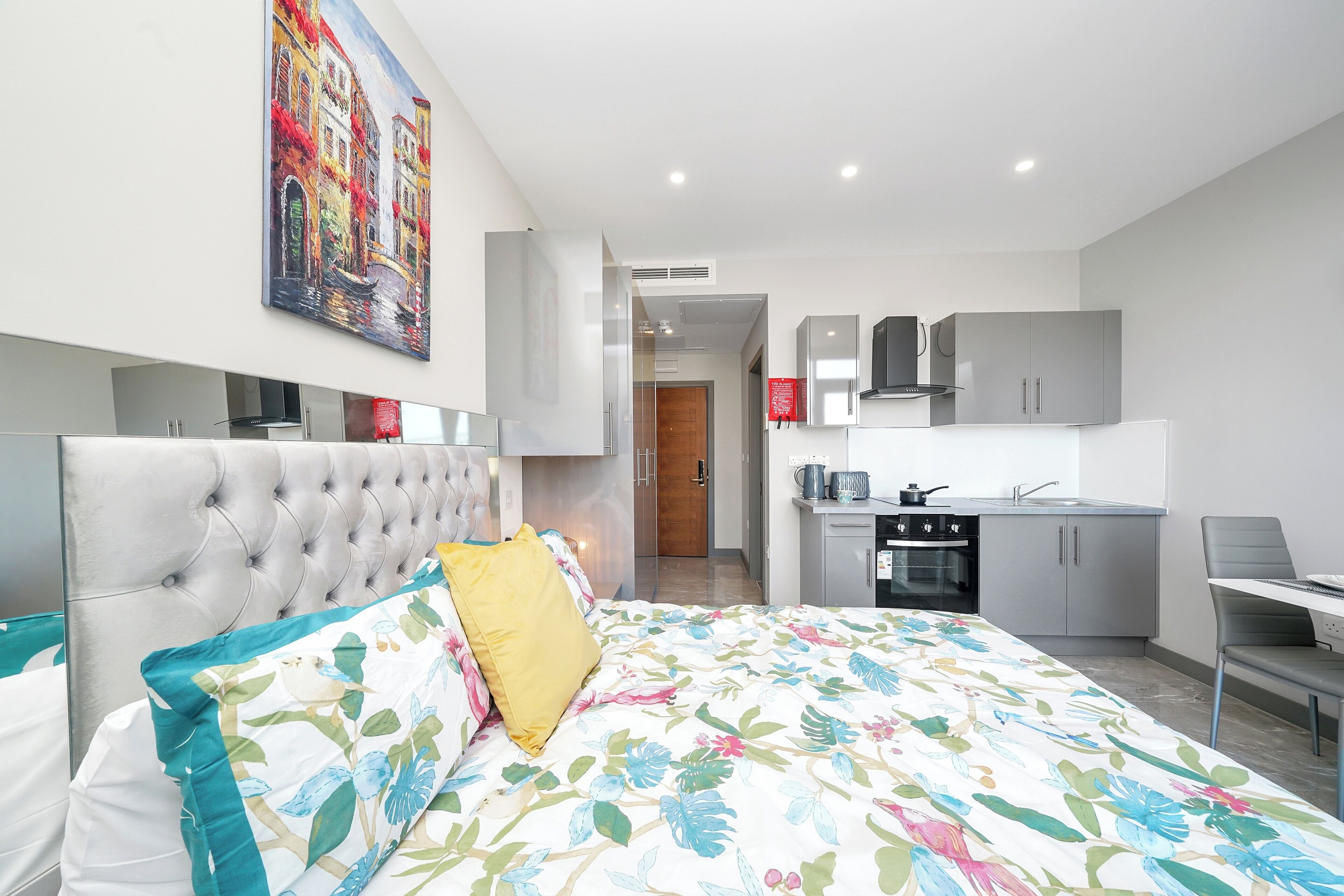 A bedroom with a tufted headboard, floral bedspread, and a large painting above the bed. Adjacent is a kitchenette with a stove, sink, and cabinets.