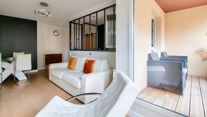A modern living room with a white sofa, a dining area with a white table, and a balcony with wicker furniture. - 123home- Suite & spa XL (Montevrain)