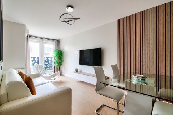 A modern living room with a sofa, a glass dining table, and a TV mounted on the wall. - 123home- Suite & spa XL (Montevrain)
