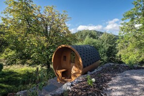 A cylindrical wooden structure with a door and a small window, situated in a natural setting with trees and a clear sky.