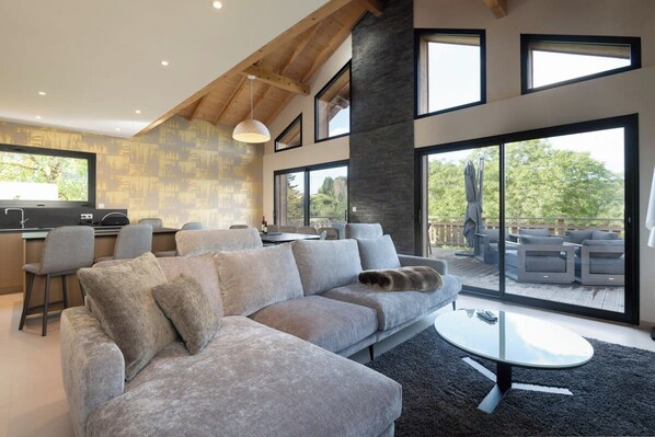 A modern living space with a sectional sofa, a dining area, and large windows. - Chalet Lac ⭐ Covered Hot Tub • Sauna • Mountain Views • Gérardmer (Gérardmer)