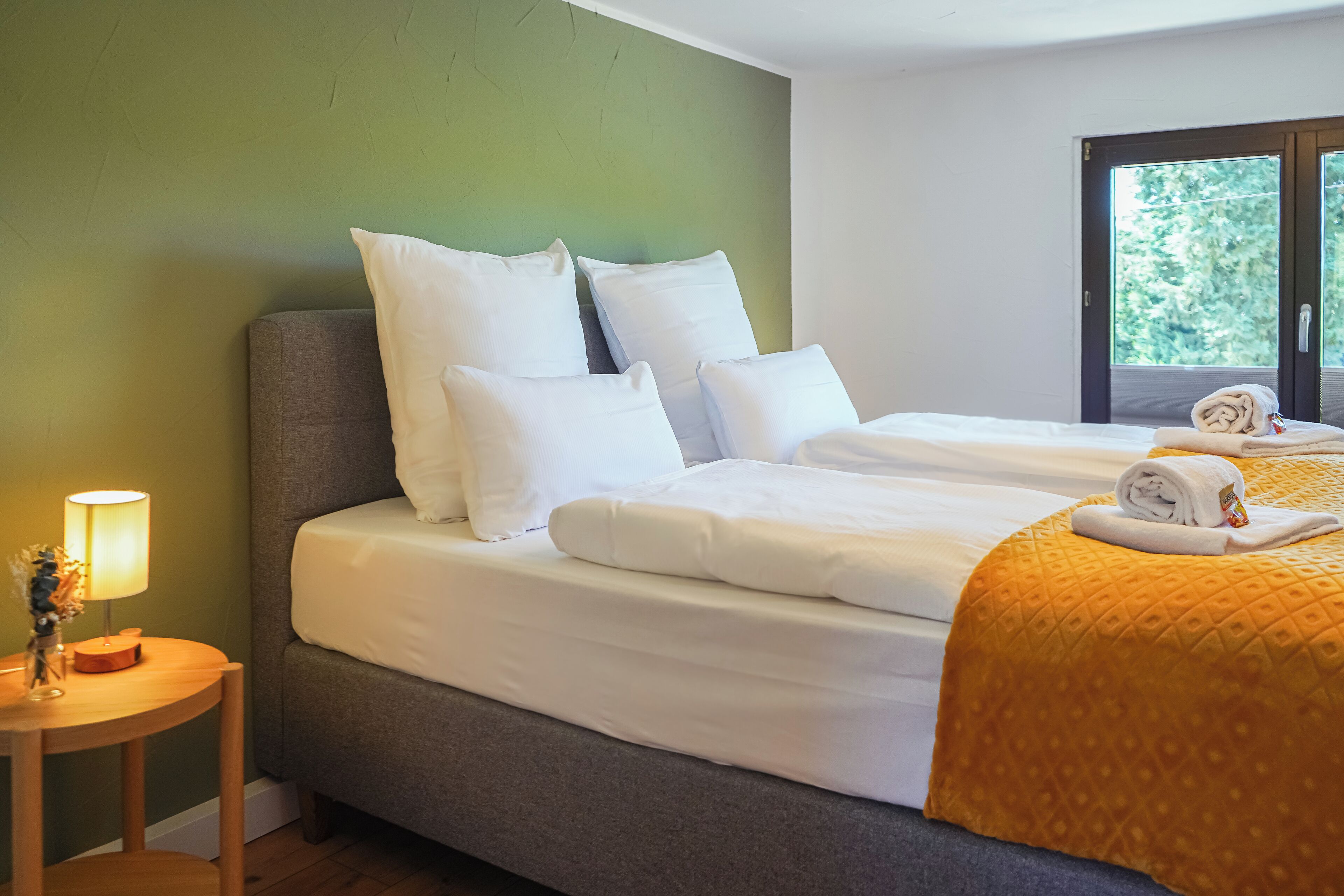 Une chambre d’hôtel avec deux lits, un mur vert, une table de chevet en bois avec une lampe, et une fenêtre donnant sur des arbres.