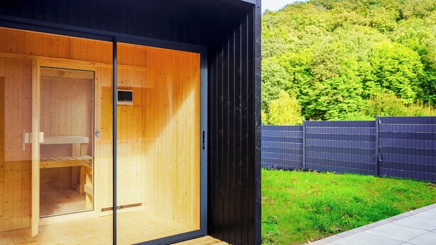 A modern wooden sauna with a glass door, a small bench inside, and a view of a green hillside.