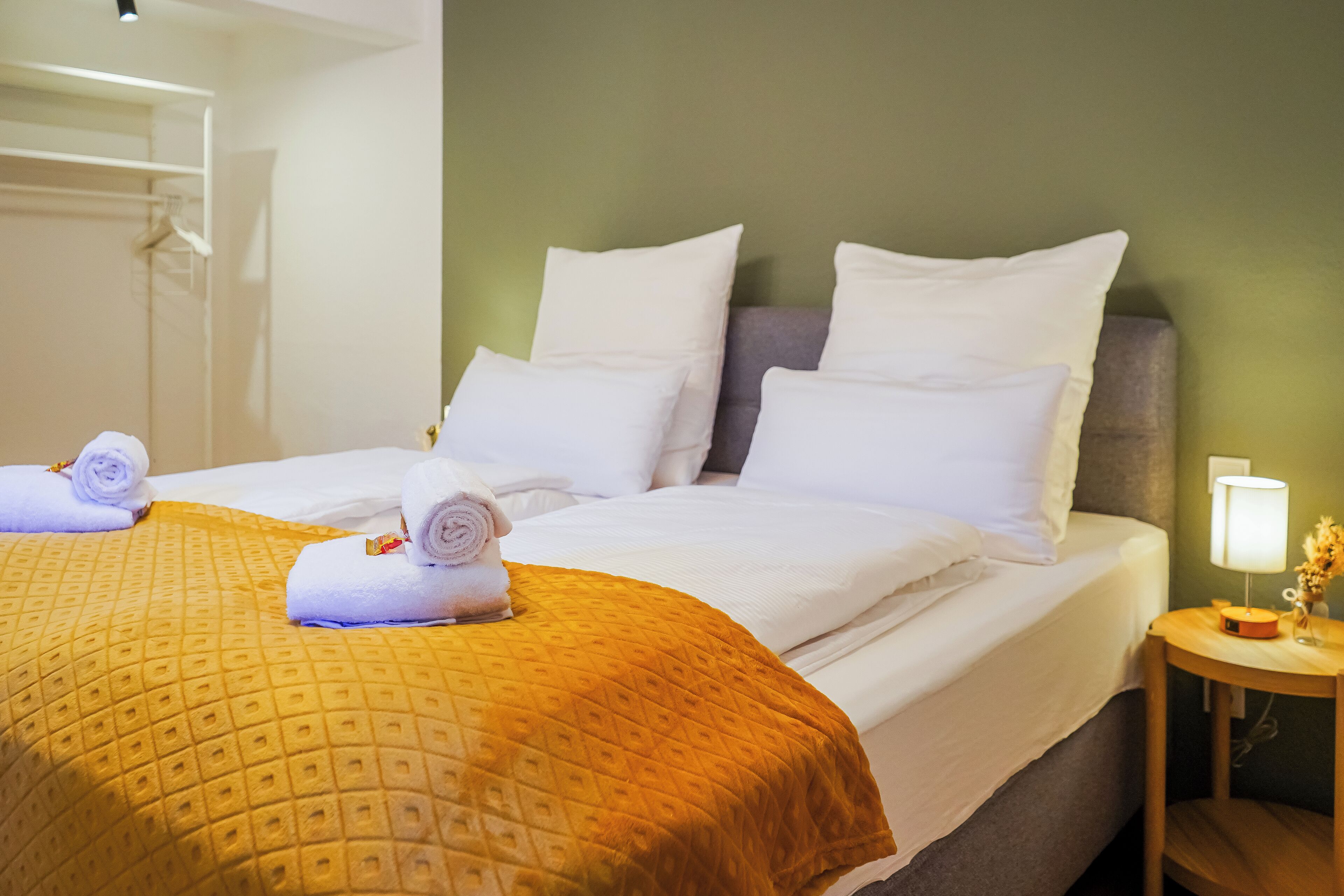 Habitación de hotel con cama, almohadas, mesita de noche con lámpara y luz en la pared.