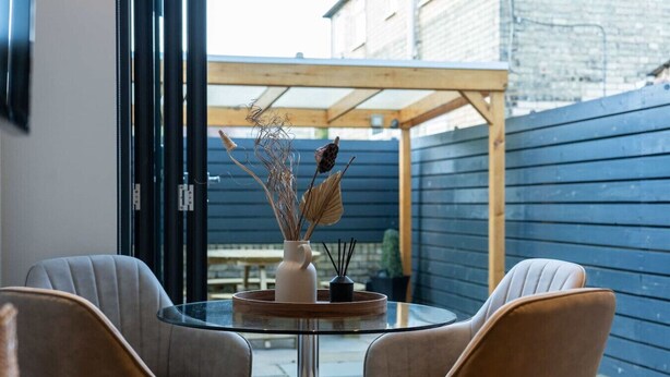 A glass table with two chairs, a vase with dried plants, and a wooden pergola in an outdoor setting.