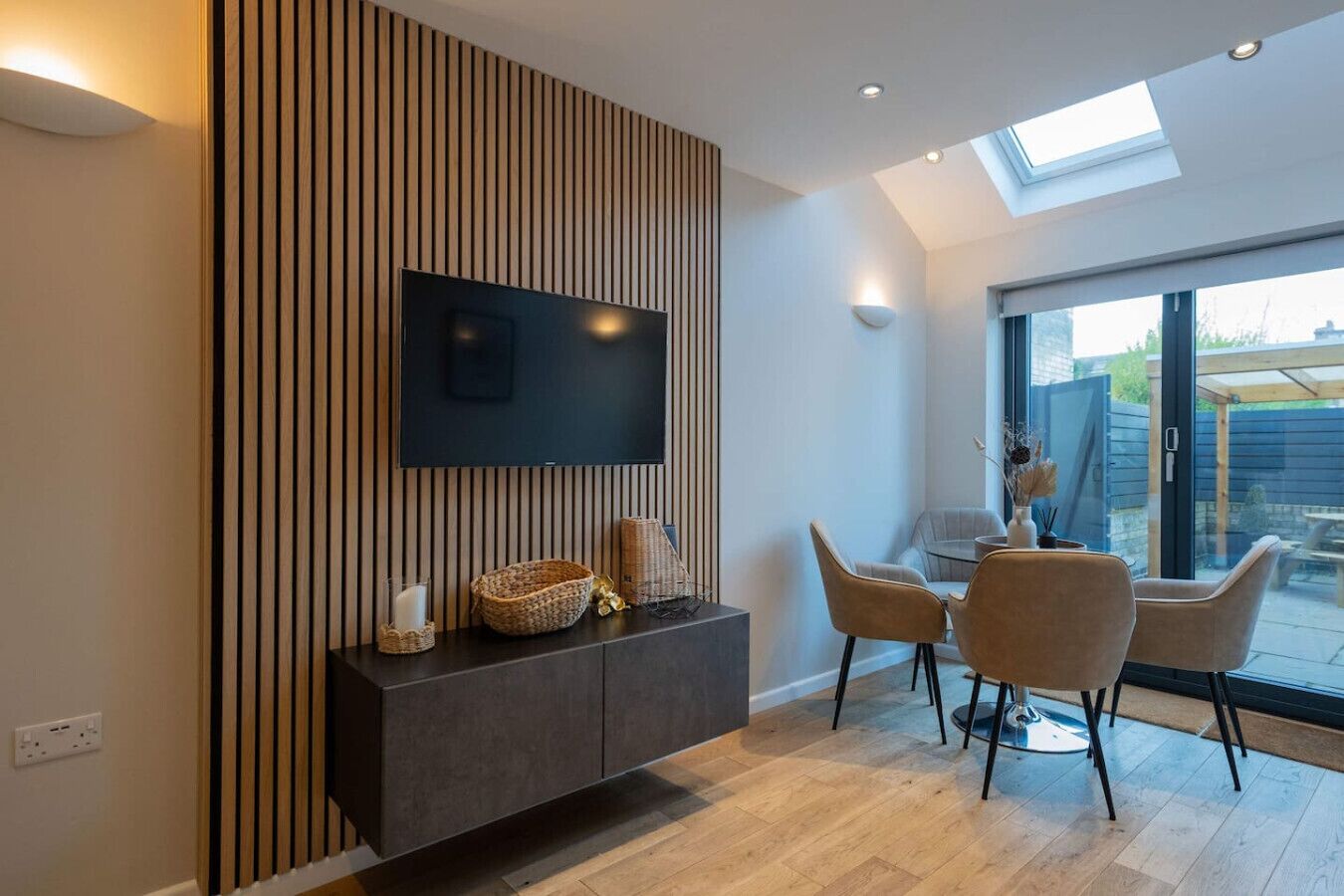 A modern living room with a flat-screen TV mounted on a wall, a dark wooden cabinet below, and a dining area with two chairs and a table.