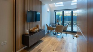 A modern living room with a flat-screen TV, wooden panel wall, and a dining area with chairs and a table.