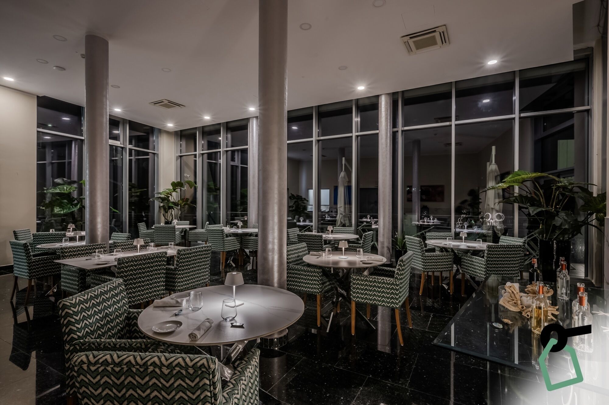 Un restaurante moderno con mesas redondas y sillas estampadas en verde, ventanas amplias y plantas de interior.