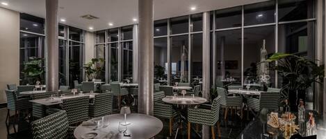Un restaurante moderno con mesas redondas y sillas estampadas en verde, ventanas amplias y plantas de interior.