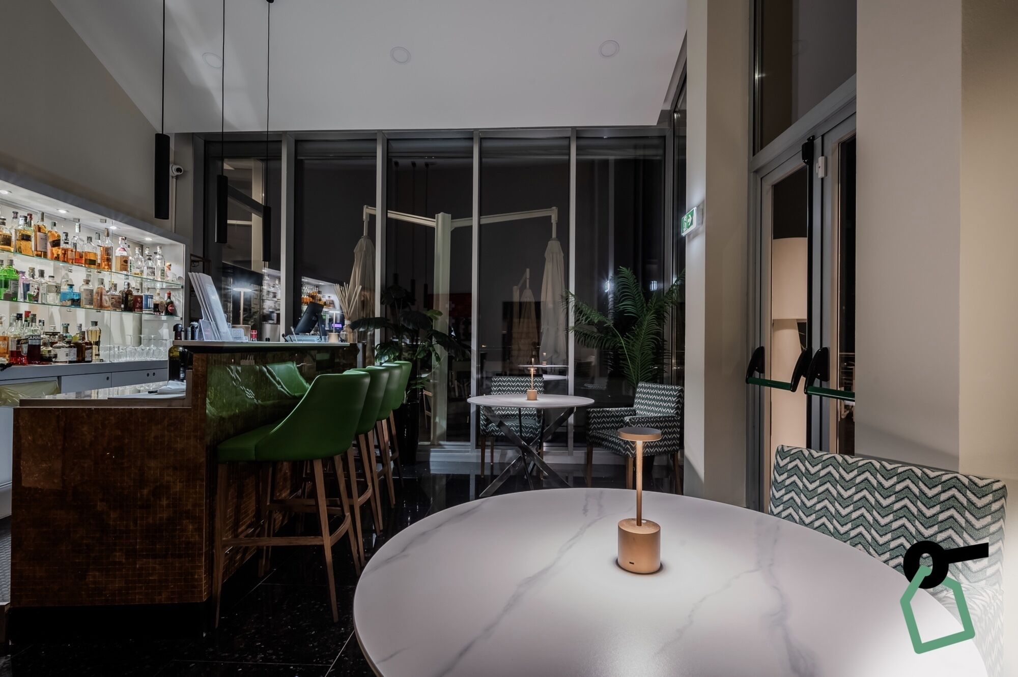 Uma área de bar moderna com um expositor de vidro cheio de garrafas, banquetas de bar verdes e uma mesa redonda com uma vela.