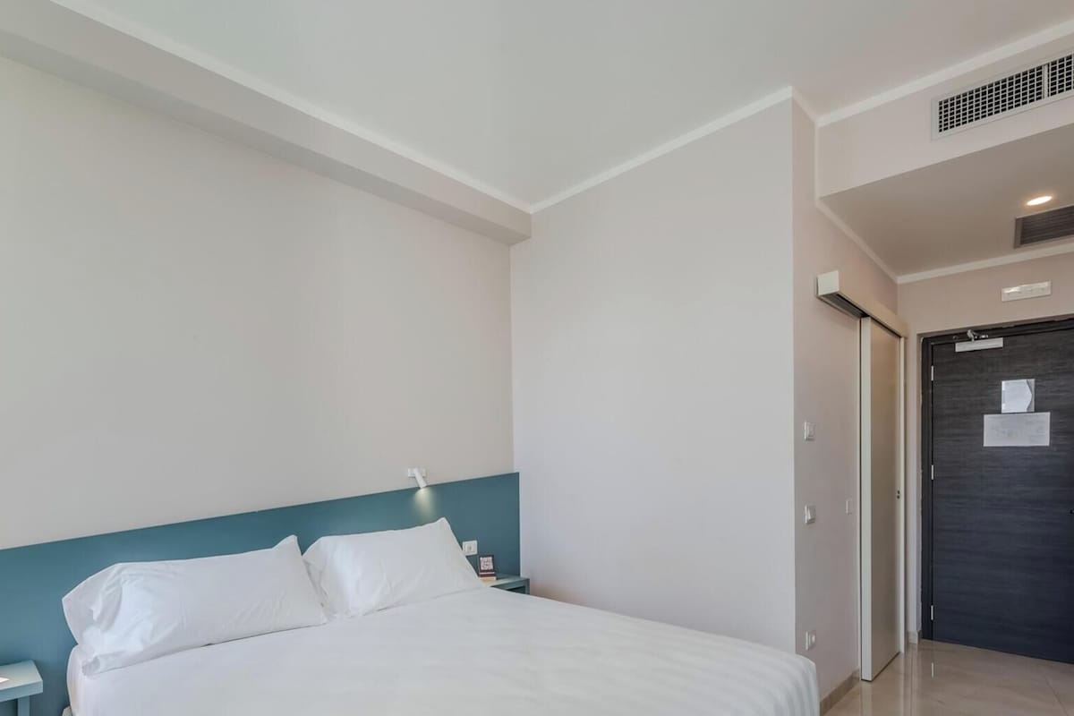 A modern hotel room with a bed, a bedside table with a phone, a wall-mounted light, and a door with a key in the lock.