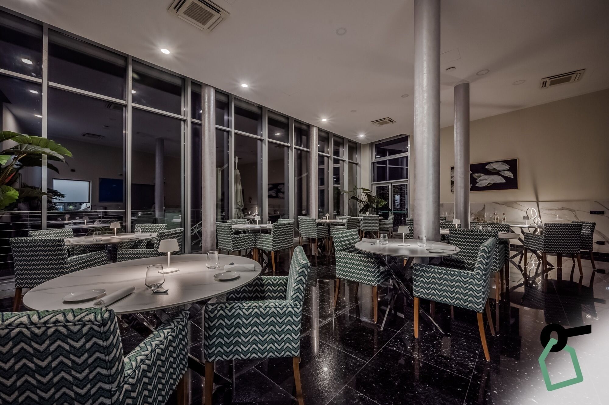 A modern dining area with round tables and zigzag patterned chairs, large windows, and a central column.