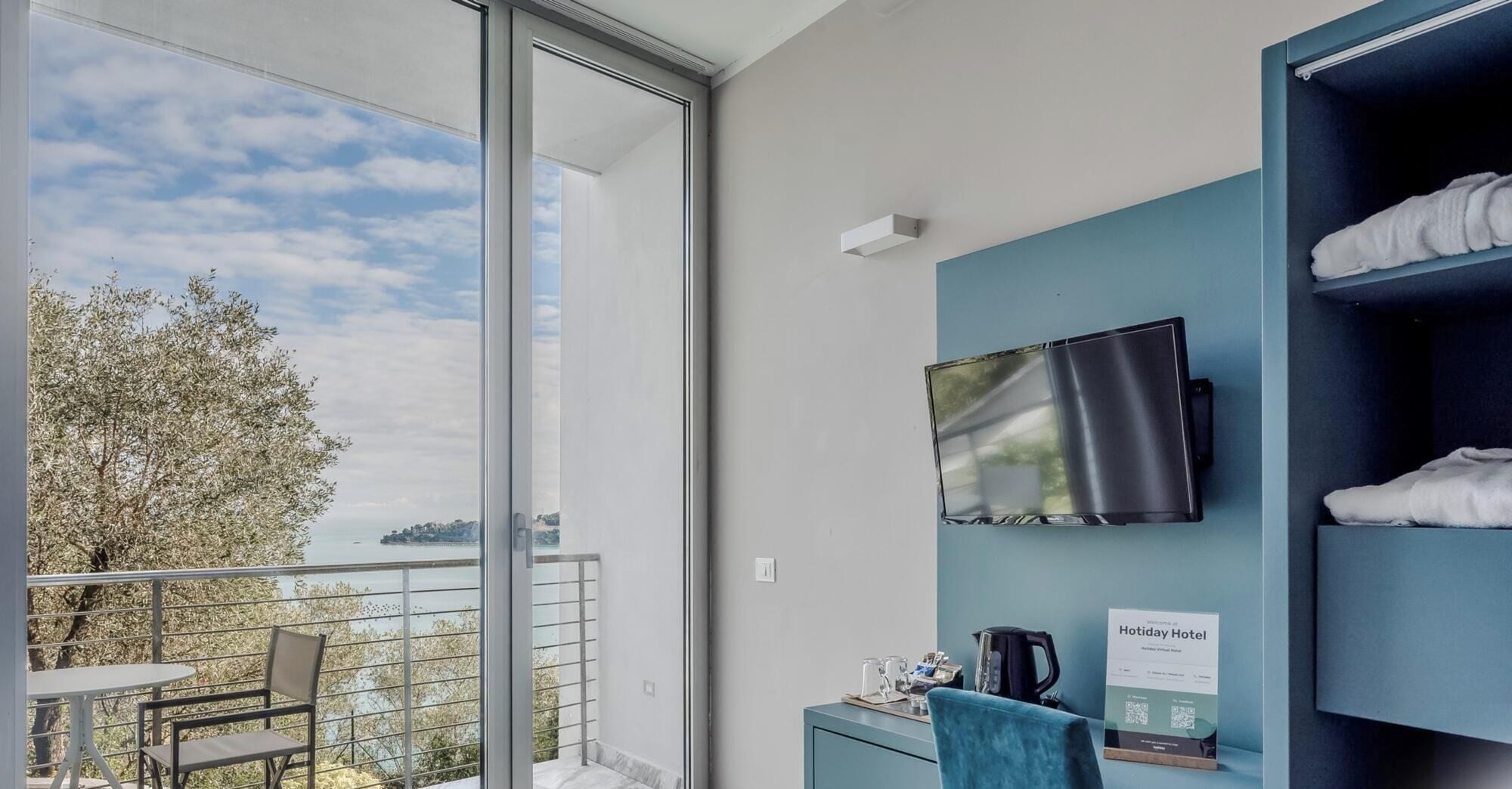 Quarto de hotel com varanda, televisão de tela plana na parede, escrivaninha com chaleira e placa indicando o tipo de quarto.