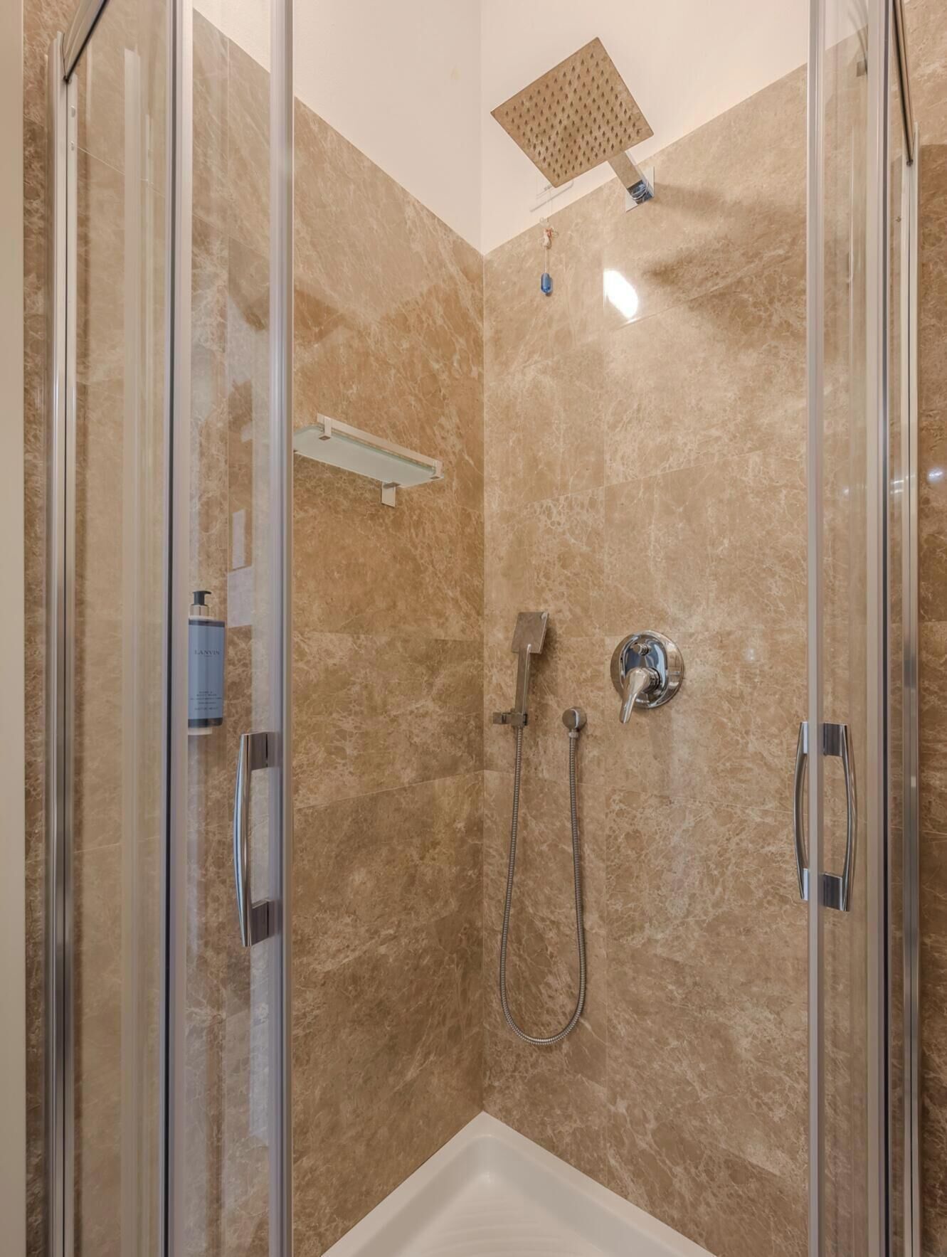 Un área de ducha con mampara de vidrio, cabezal de ducha manual y grifo empotrado en la pared.