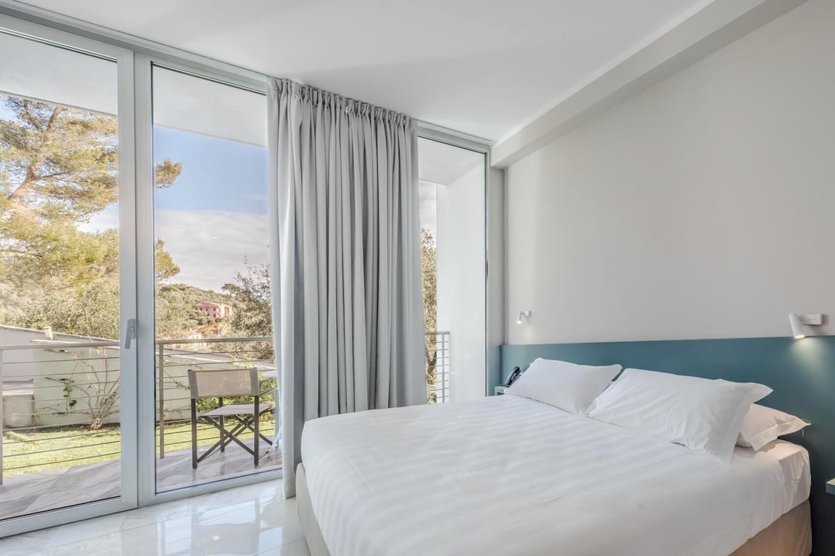 A modern hotel room with a large bed, a nightstand, and a view of a balcony with outdoor furniture.