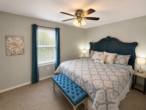 Quarto com cama, um banquinho, ventoinha no teto e janela com cortinas.