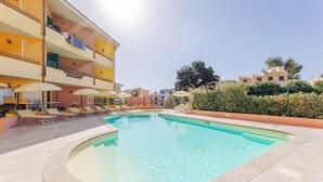 A swimming pool with lounge chairs and umbrellas, surrounded by a multi-story building with balconies. - Apartments i Mirti Bianchi (Santa Teresa di Gallura)