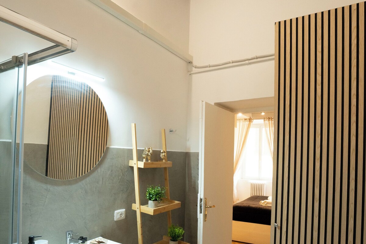 A modern bathroom with a large mirror, a sink, and a shower. There is a wooden shelf with plants and towels.