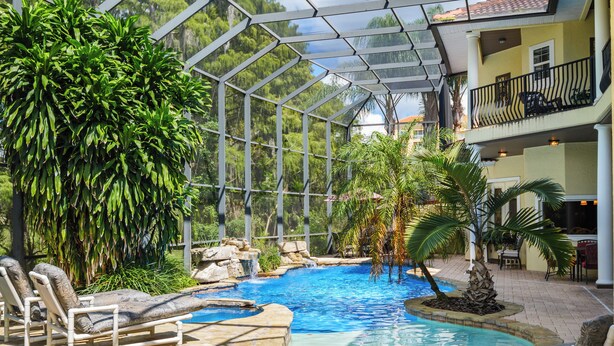 A pool area with lounge chairs, a glass-enclosed structure, and tropical plants. - Magnificent 8 Bed 8 Bath Custom  6500+ SqFt. Pool/spa. NEW DISCOUNTED 2026 rates (Kissimmee)