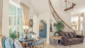 A spacious living room with a blue sofa, a wooden coffee table, and a chandelier. - Magnificent 8 Bed 8 Bath Custom  6500+ SqFt. Pool/spa. NEW DISCOUNTED 2026 rates (Kissimmee)