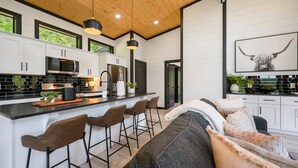 A modern kitchen with white cabinets, a central island with bar stools, and a dining area with a sofa and cushions. - Stargaze from Your Private Hot Tub (Broken Bow)