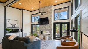 A modern living room with a stone fireplace, large windows, and a ceiling fan. - Stargaze from Your Private Hot Tub (Broken Bow)