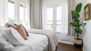 Un dormitorio con cama, cortinas blancas, un ventanal grande y una planta en maceta.