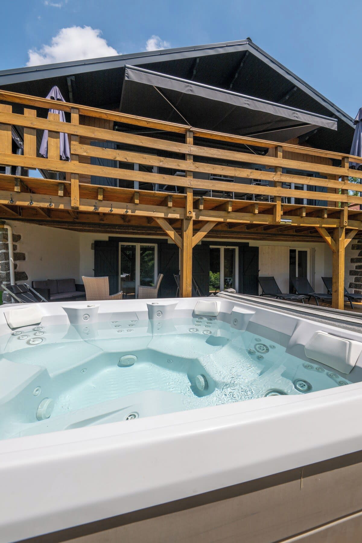 A hot tub with a wooden deck and lounge chairs in front, under a covered area with a view of a building.