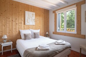 Un dormitorio con pared de paneles de madera, una cama grande con ropa blanca, una ventana con vista a la vegetación y una mesita de noche con una lámpara.