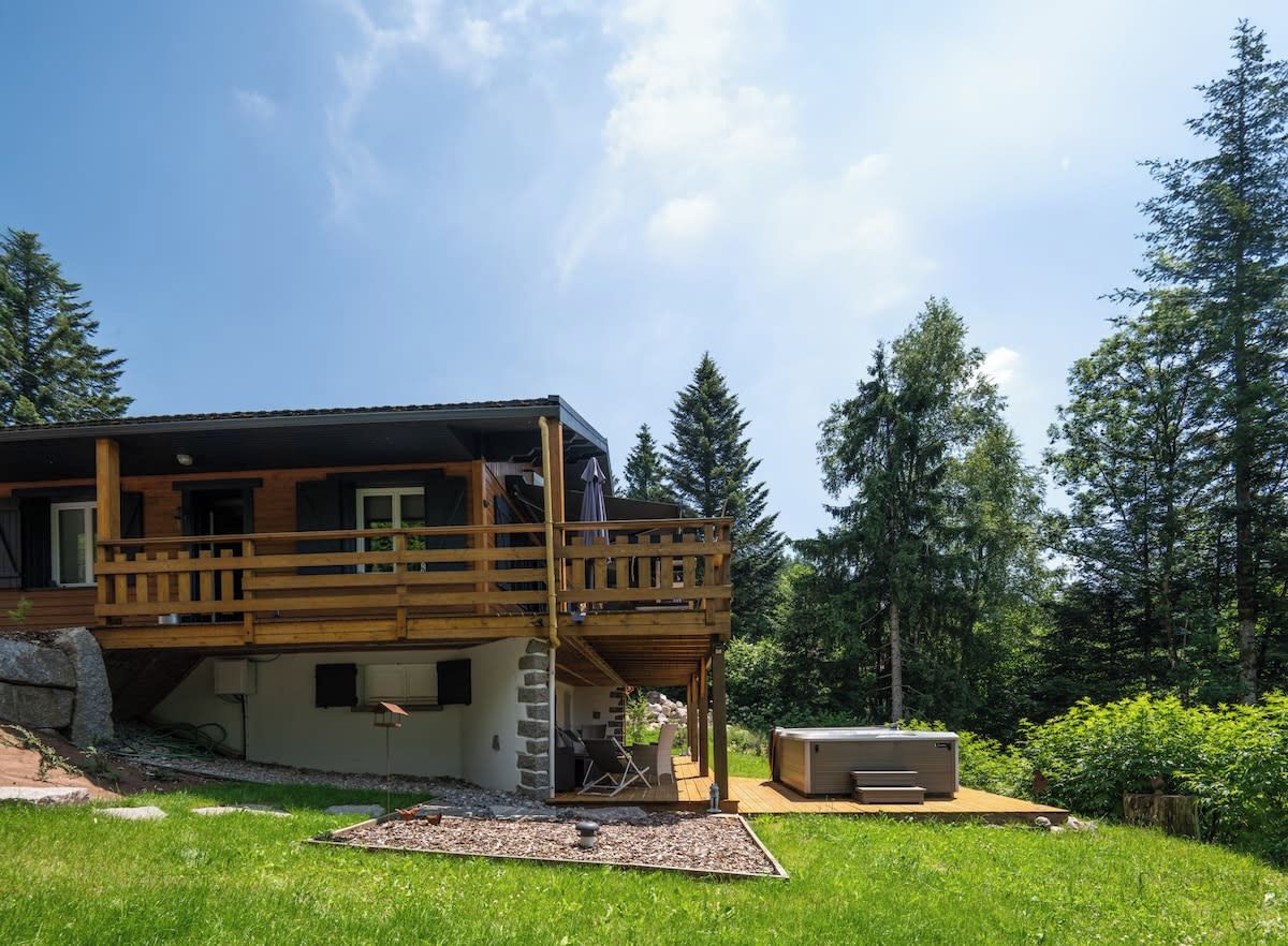 A two-story wooden cabin with a balcony, surrounded by a grassy yard and trees.