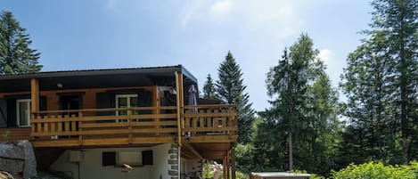 A two-story wooden cabin with a balcony, surrounded by a grassy yard and trees.
