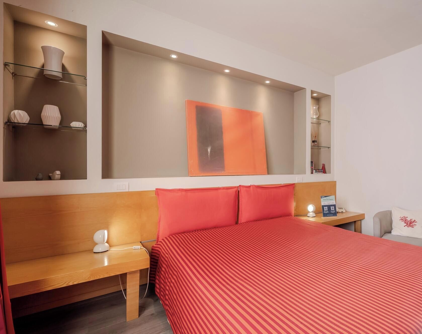A hotel room with a large bed, a bedside table, a lamp, a mirror, and a shelf with decorative items.