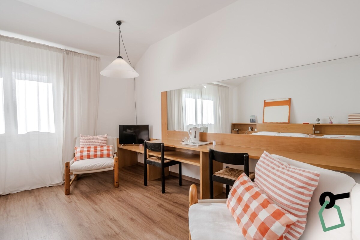 Quarto de hotel com escrivaninha de madeira, uma cadeira, uma cama, televisão e espelho.