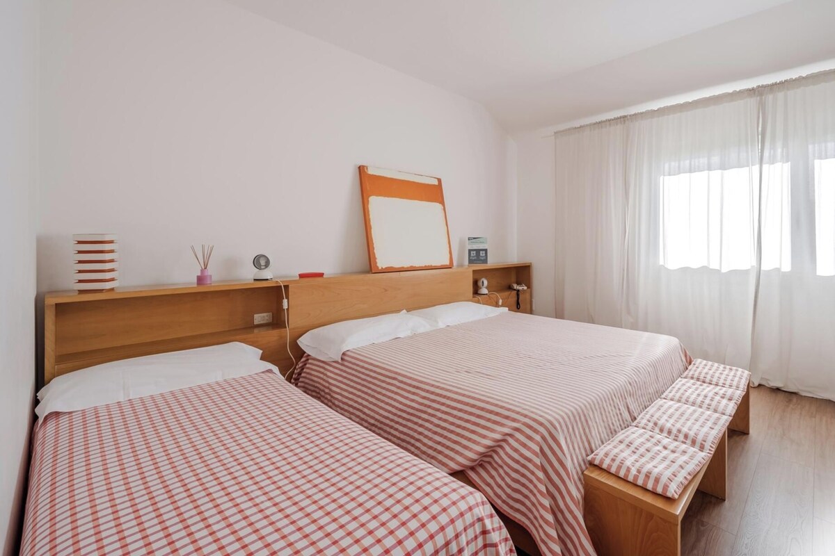A hotel room with two beds, a bedside table with a lamp, a mirror, and a window with curtains.