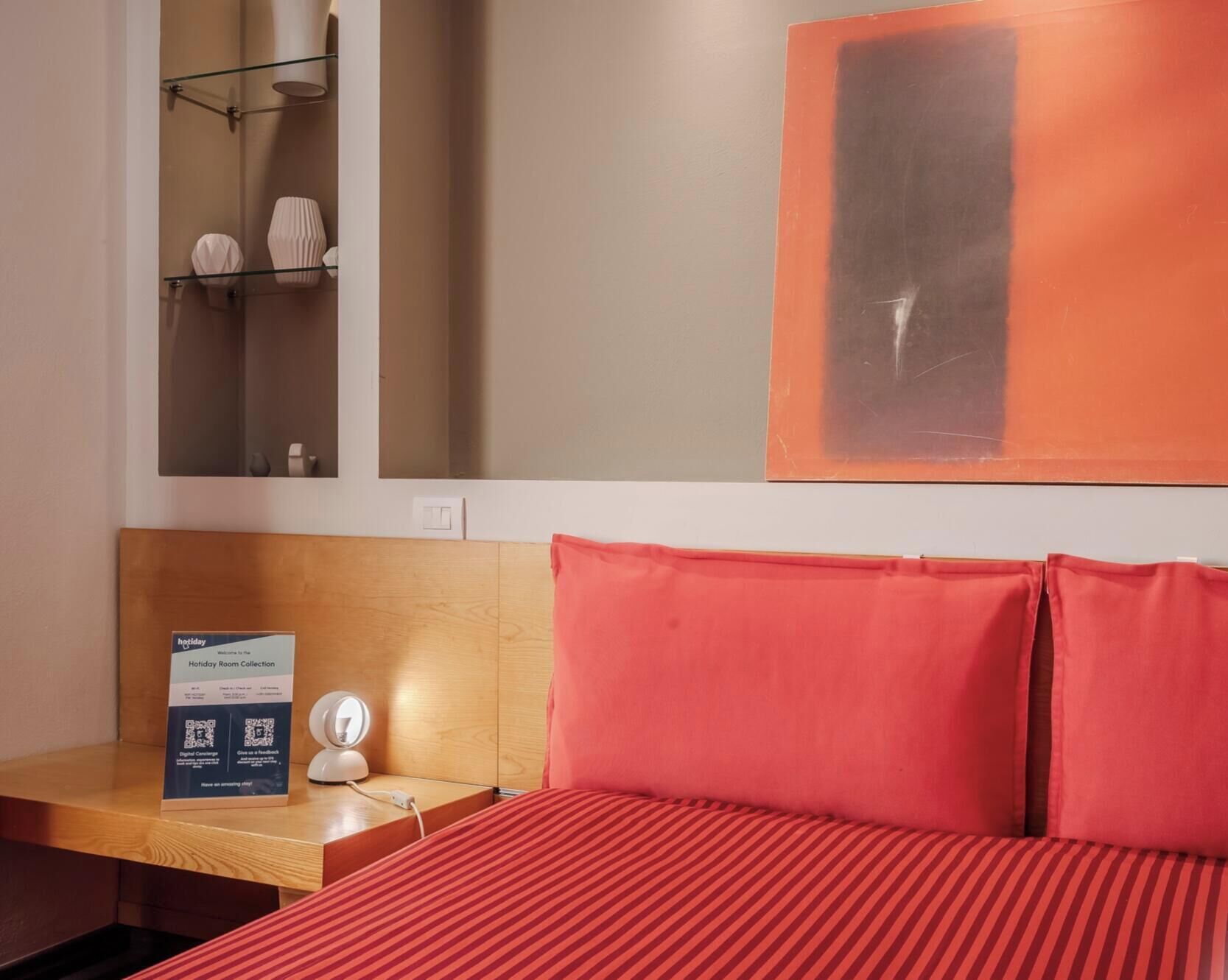 Ein Hotelzimmer mit Bett, Nachttisch, digitaler Uhr, Schild für die Holiday-Room-Kollektion und einem Spiegel mit Ablageflächen darüber.
