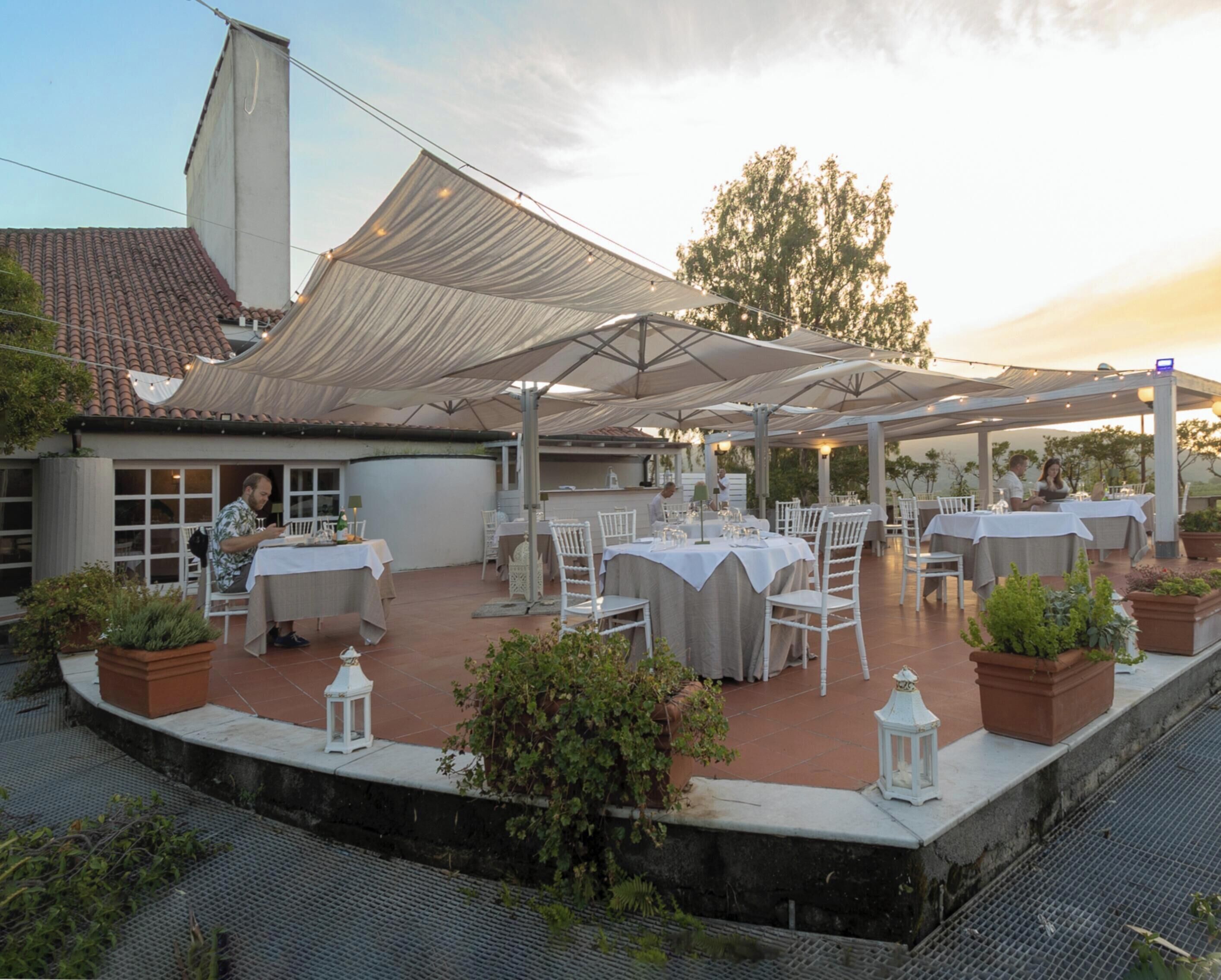 Un área de comedor al aire libre con mesas y sillas, cubierta por un toldo grande y rodeada de plantas en macetas.