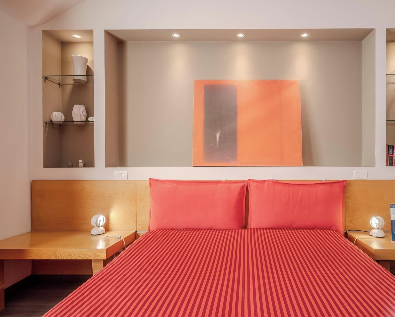 Camera d'albergo con un letto grande, due comodini, una mensola a parete con oggetti decorativi e un grande quadro astratto.