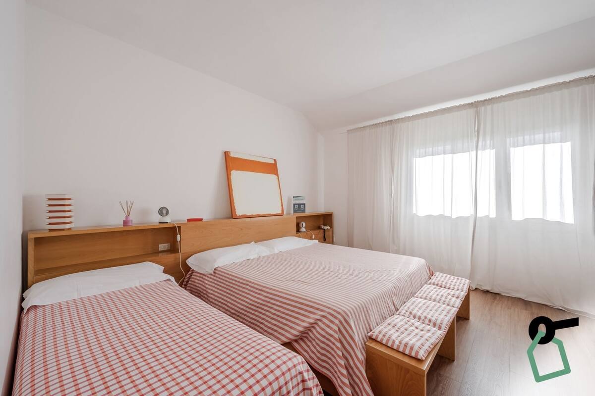 A hotel room with two beds, a wooden headboard, a bedside table, a mirror, a window with sheer curtains, and a small shelf with items.