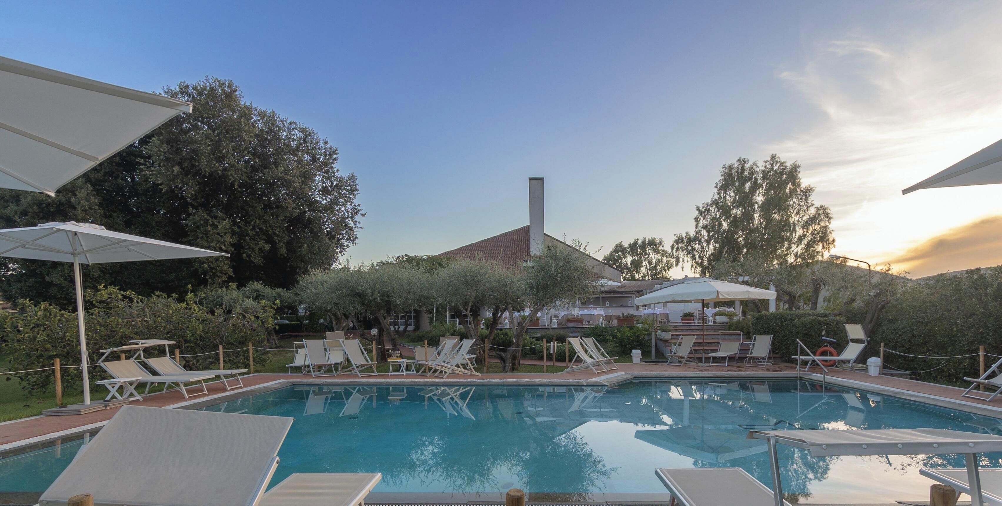 Una piscina amb gandules i ombrelles, envoltada d'arbres i un edifici amb una xemeneia.