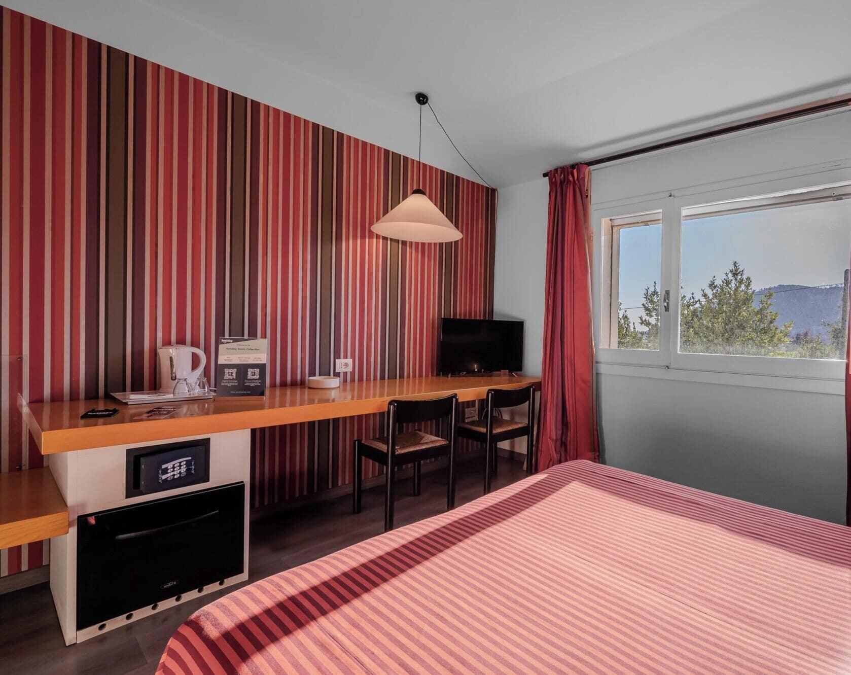 A hotel room with a bed, a desk with a television, a coffee maker, and a view of the outdoors.
