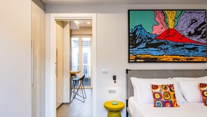 Une chambre d’hôtel moderne avec un lit, des coussins colorés, un tabouret jaune et un tableau éclatant représentant un volcan.